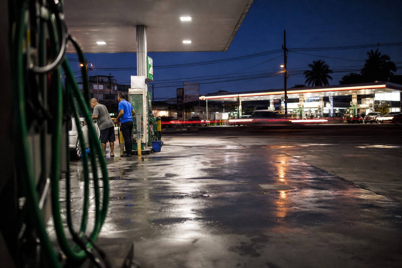 Dois postos de combustível concorrentes iluminados em lados opostos de uma avenida ao anoitecer