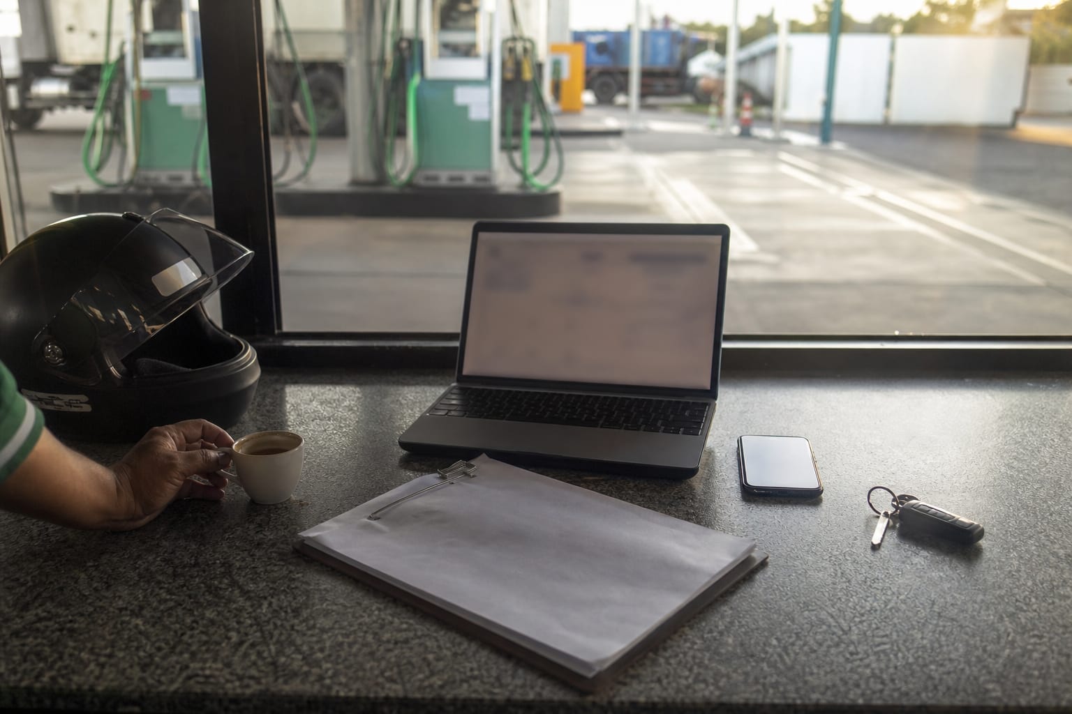 Mesa de escritório de posto com notebook, celular, papéis e capacete, com a pista visível pela janela